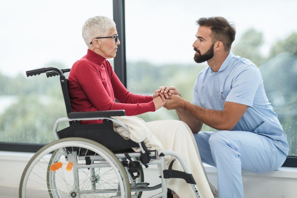 A support worker crouching to talk with a woman using a wheelchair beside a sunlit window